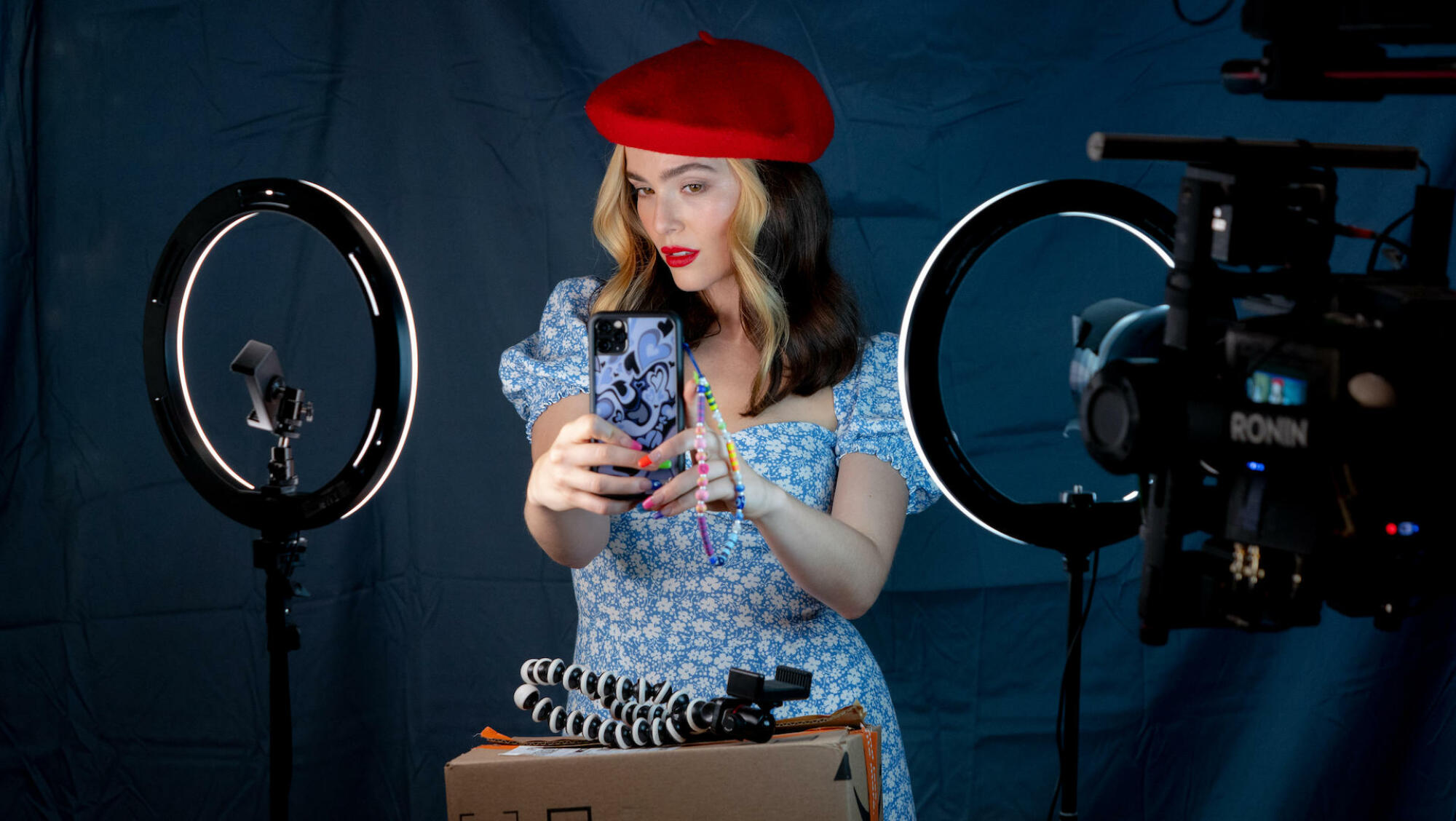 Une femme portant un béret rouge et une robe bleue prend un selfie sur son téléphone avec des anneaux lumineux.