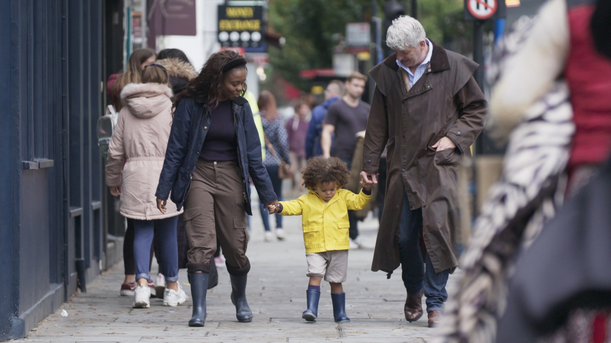 Un homme et une femme tiennent les mains d'un petit enfant marchant entre eux dans la rue.  L'enfant porte un imperméable jaune.