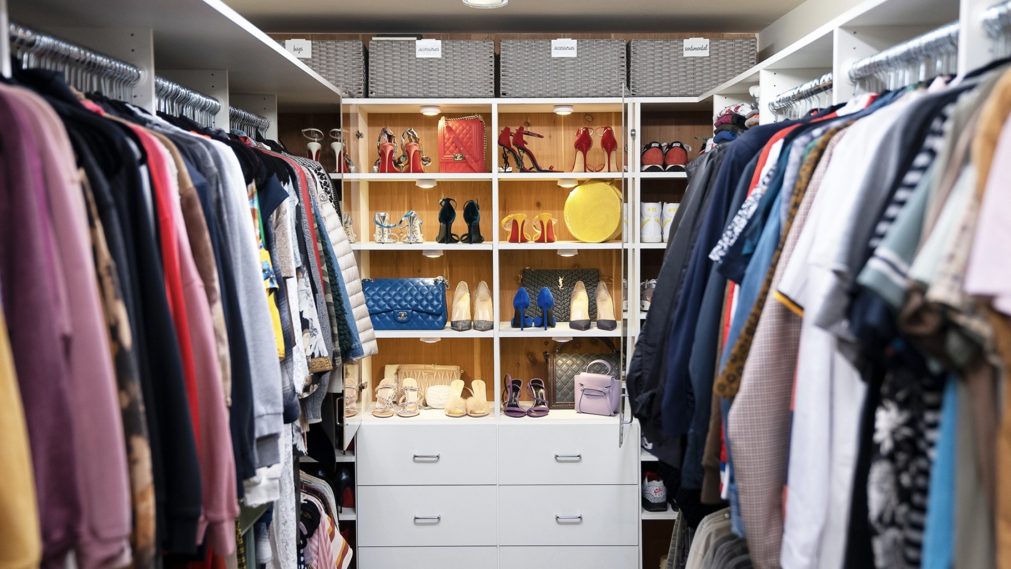 Un dressing très organisé, avec des vêtements sur des cintres sur deux murs différents et des chaussures et des sacs à main au fond de la pièce.