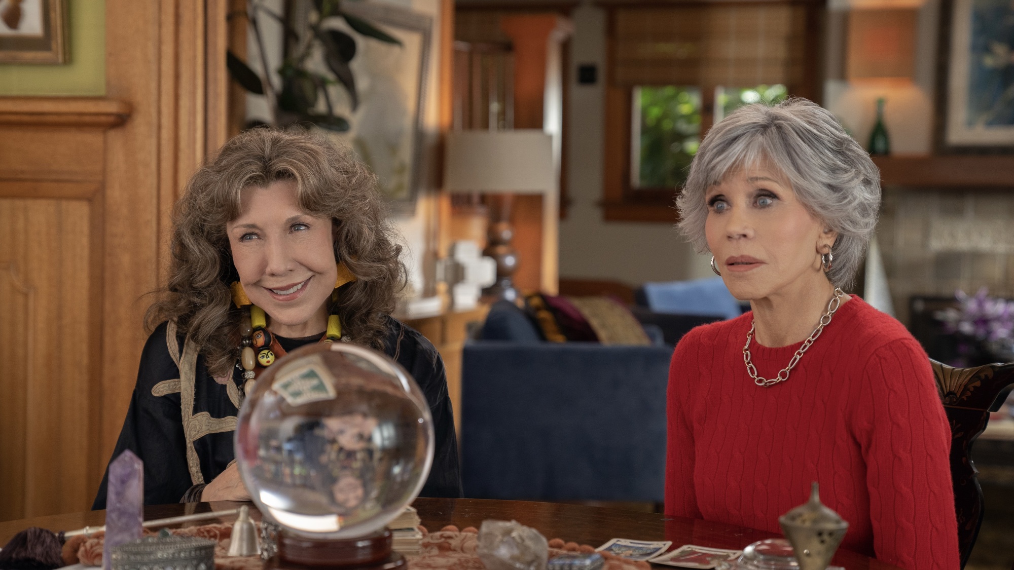 Deux femmes plus âgées, l'une en pull rouge, l'autre en chemise drapée noire, sont assises à une table avec une boule de cristal dessus.