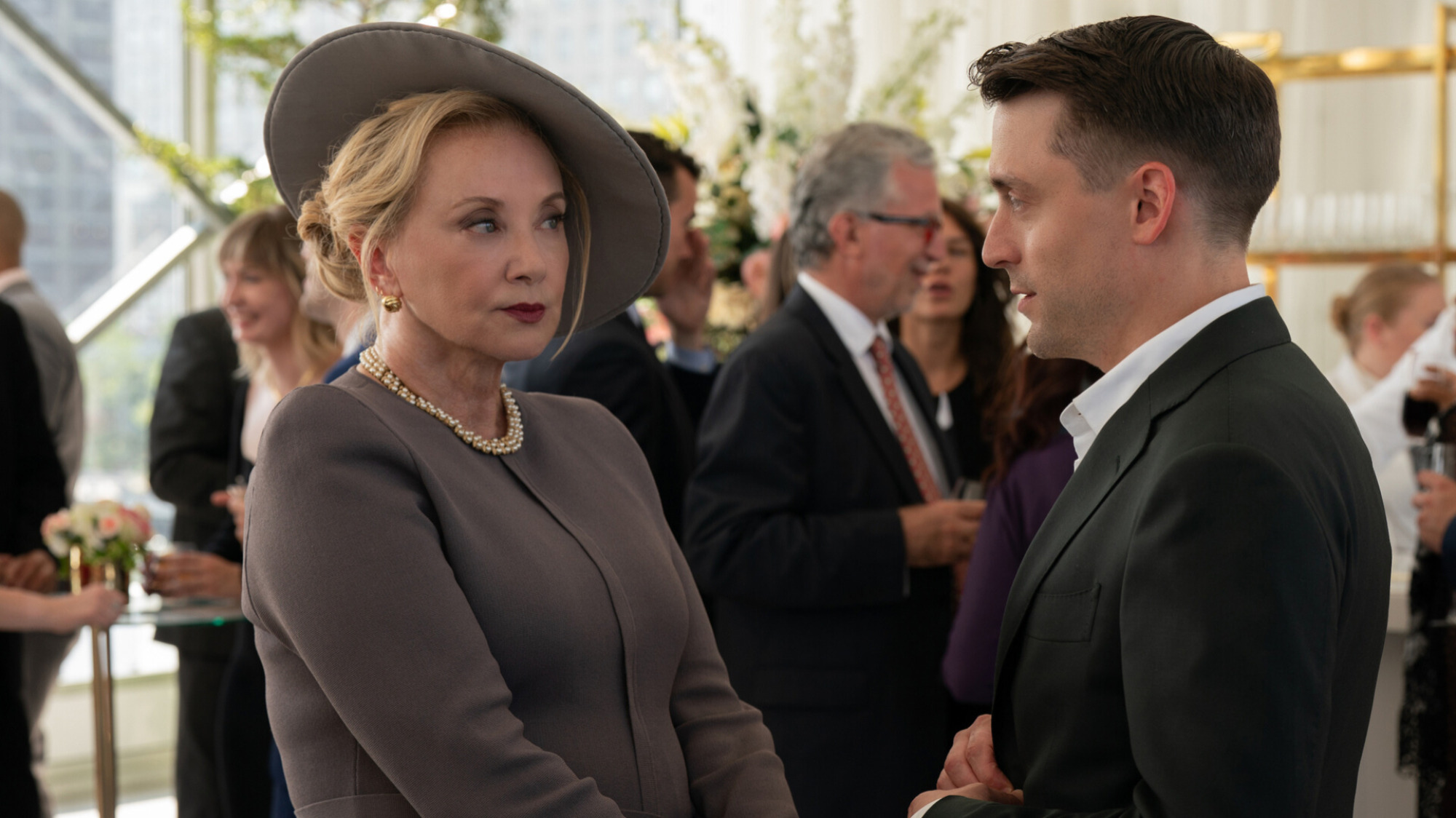 Une femme blonde portant une robe et un chapeau gris parle à un homme portant un costume.