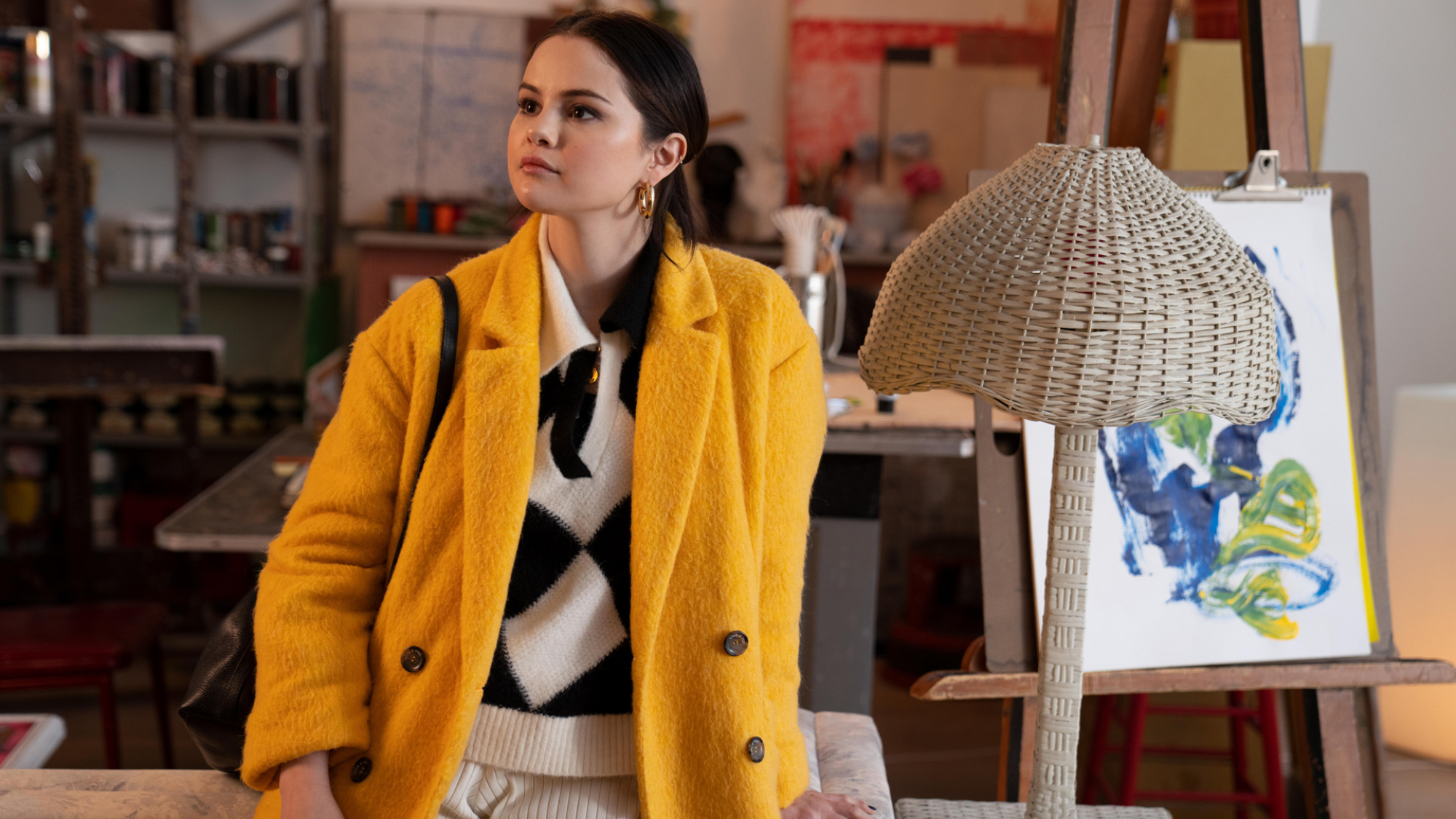 Une jeune femme en pull à carreaux noir et blanc et trench jaune s'appuie sur une table.