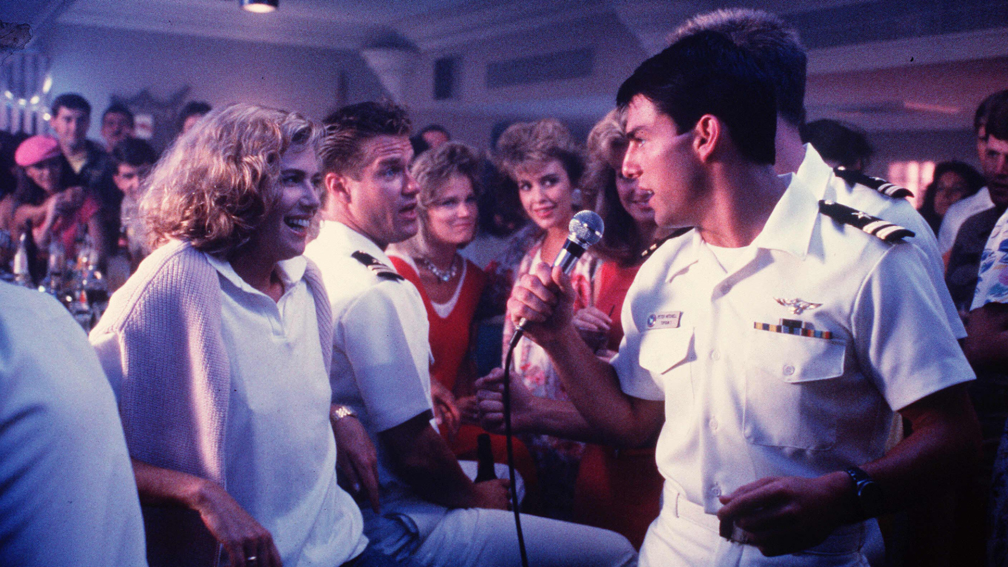 Un homme en uniforme de la marine chante à une femme dans un bar, entouré de gens.