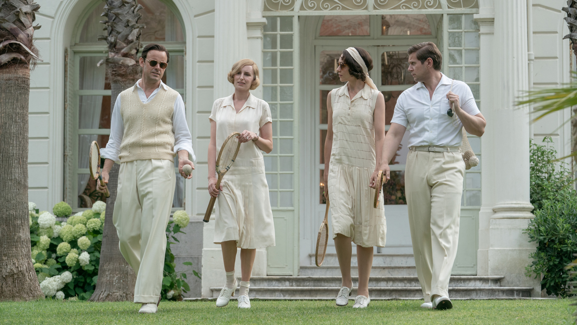 Quatre personnes en tenue de tennis des années 1930 marchent sur une pelouse.