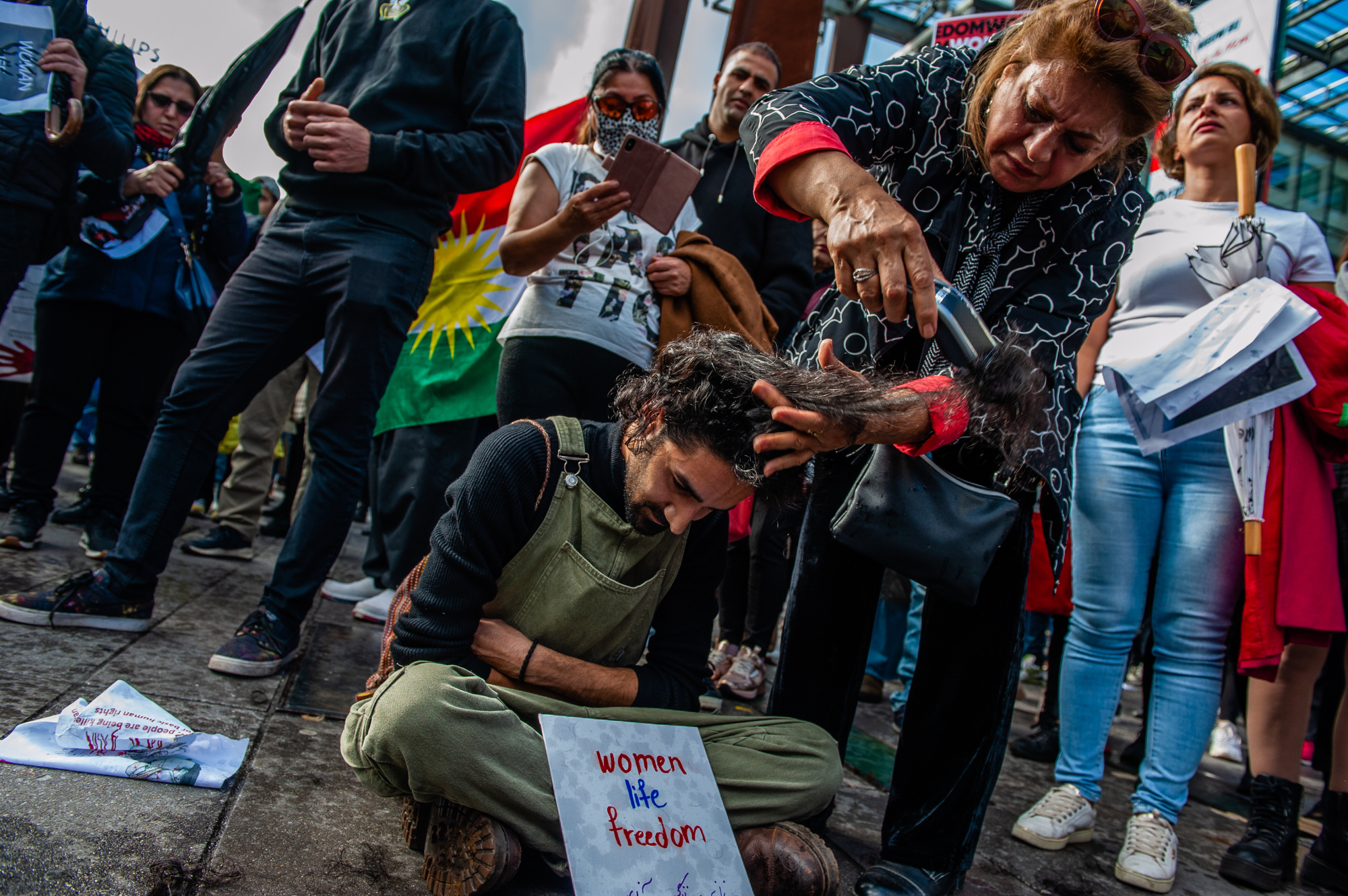 Un homme aux longs cheveux bouclés est assis sur un trottoir entouré d'une foule de gens.  Une femme âgée tient une section de ses cheveux et la coupe à l'aide d'un rasoir électrique.