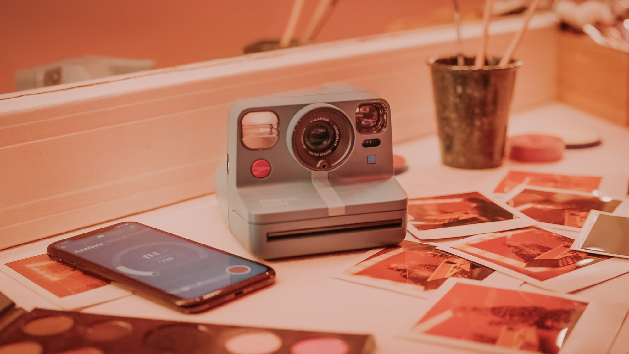 Un appareil photo instantané bleu-gris est posé sur une table parmi des tirages et un téléphone.  L'image est recouverte d'une teinte rouge.