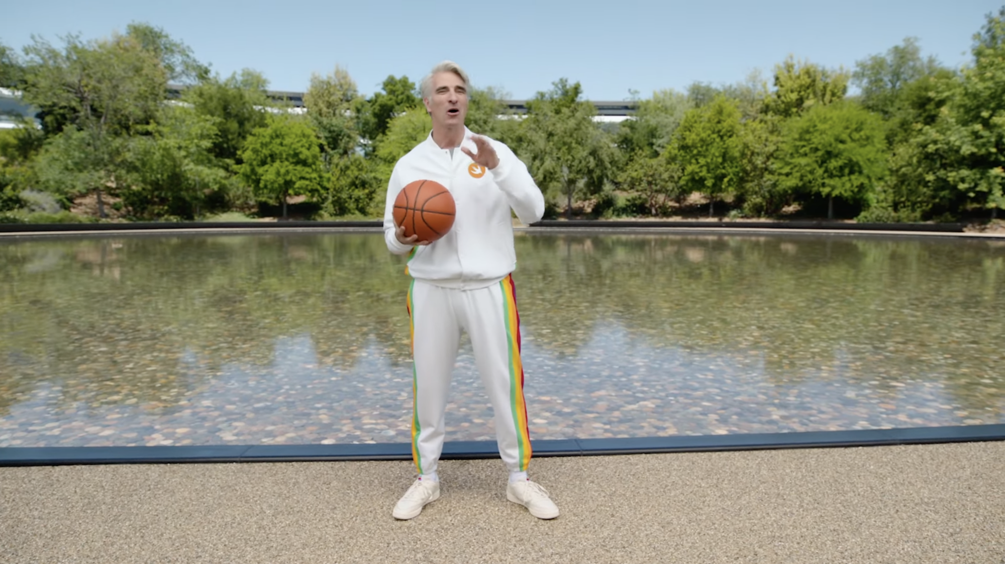 Craig Federighi tient un ballon de basket dans sa paume et porte un survêtement blanc avec des rayures multicolores sur le côté.