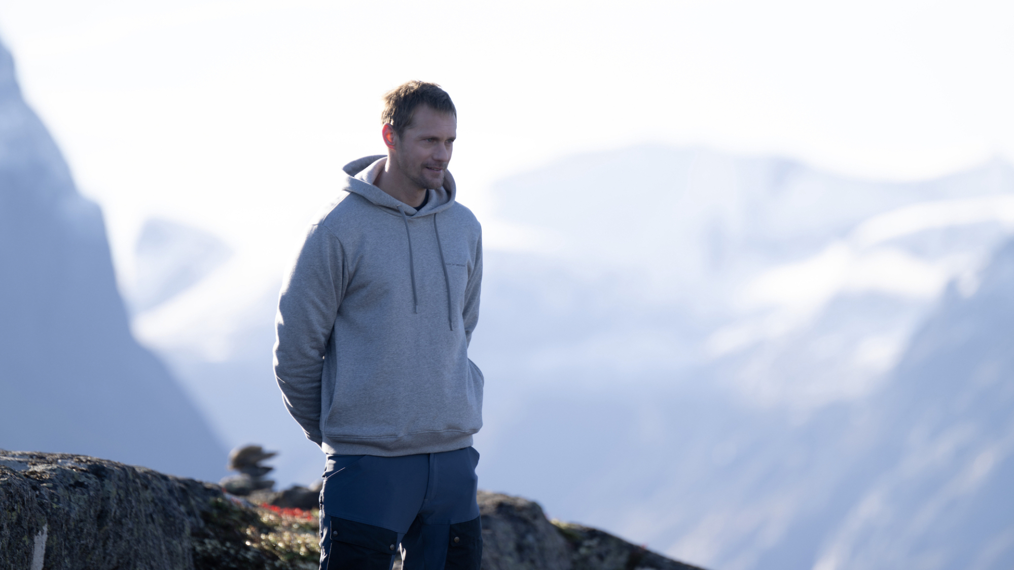 Un homme portant un sweat à capuche se tient les mains derrière le dos au sommet d'une montagne norvégienne dans l'émission télévisée 