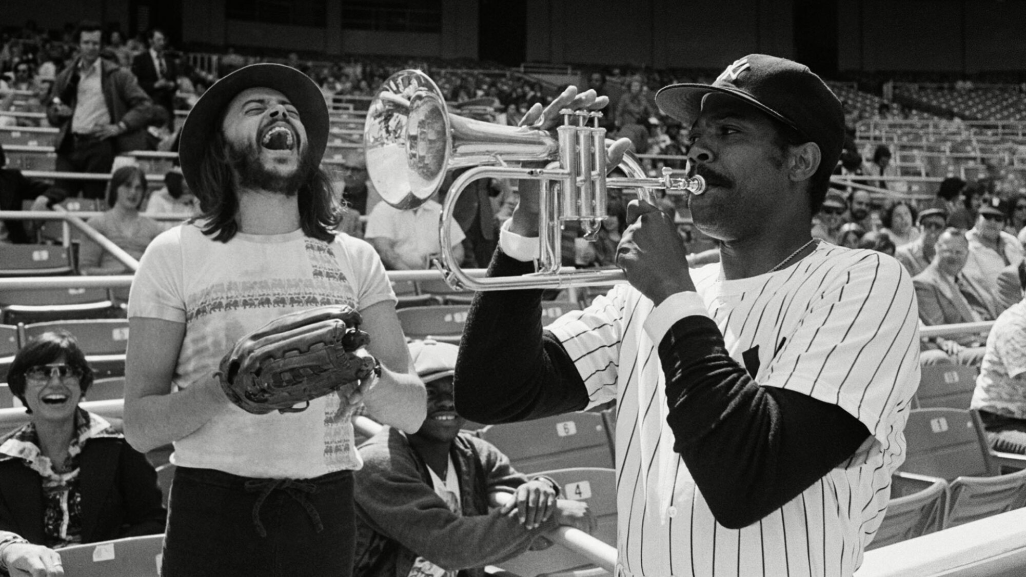 Dock Ellis, vêtu d'un uniforme des Yankees de New York, joue de la trompette tandis que le musicien de jazz Chuck Mangione se tient à proximité en riant.