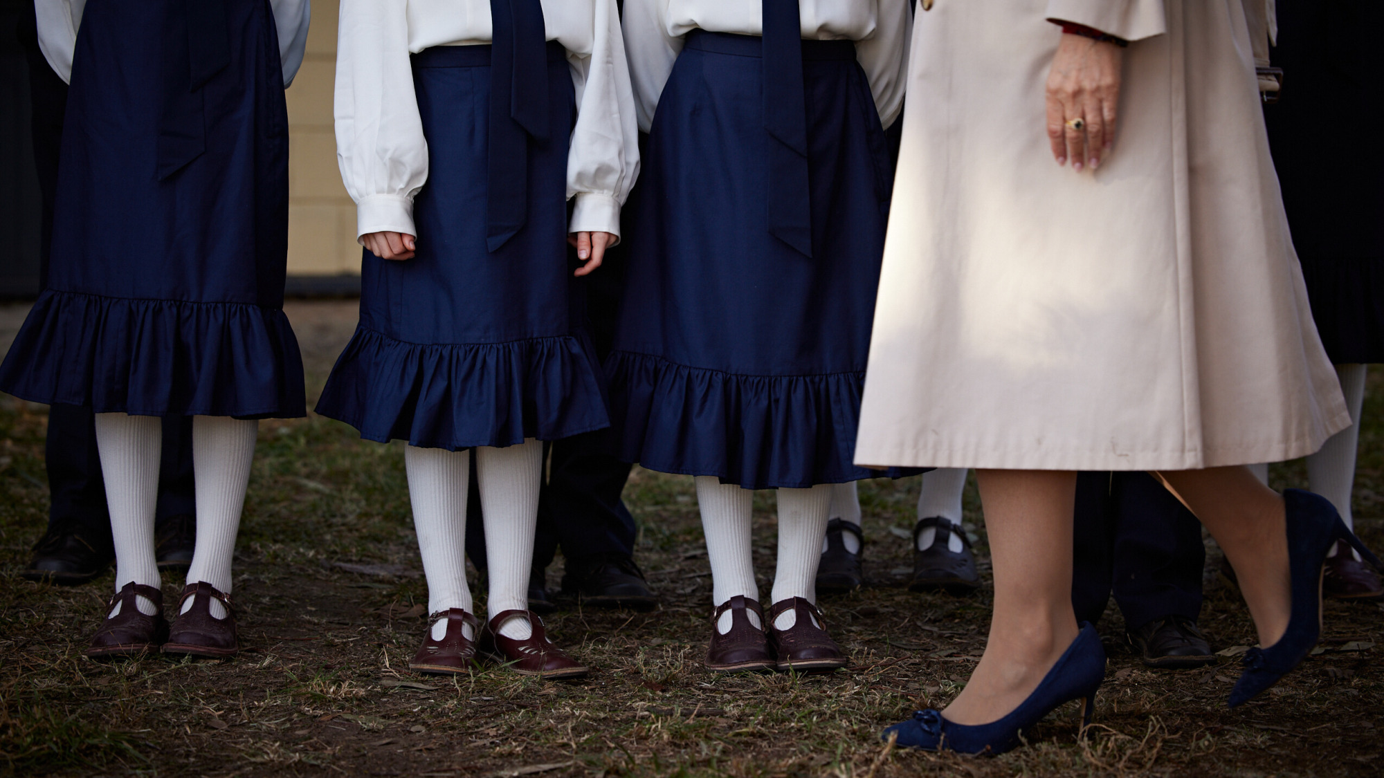 Un groupe d'enfants portant des uniformes scolaires fait la queue, tandis qu'un adulte vêtu d'une jupe rose marche devant eux.