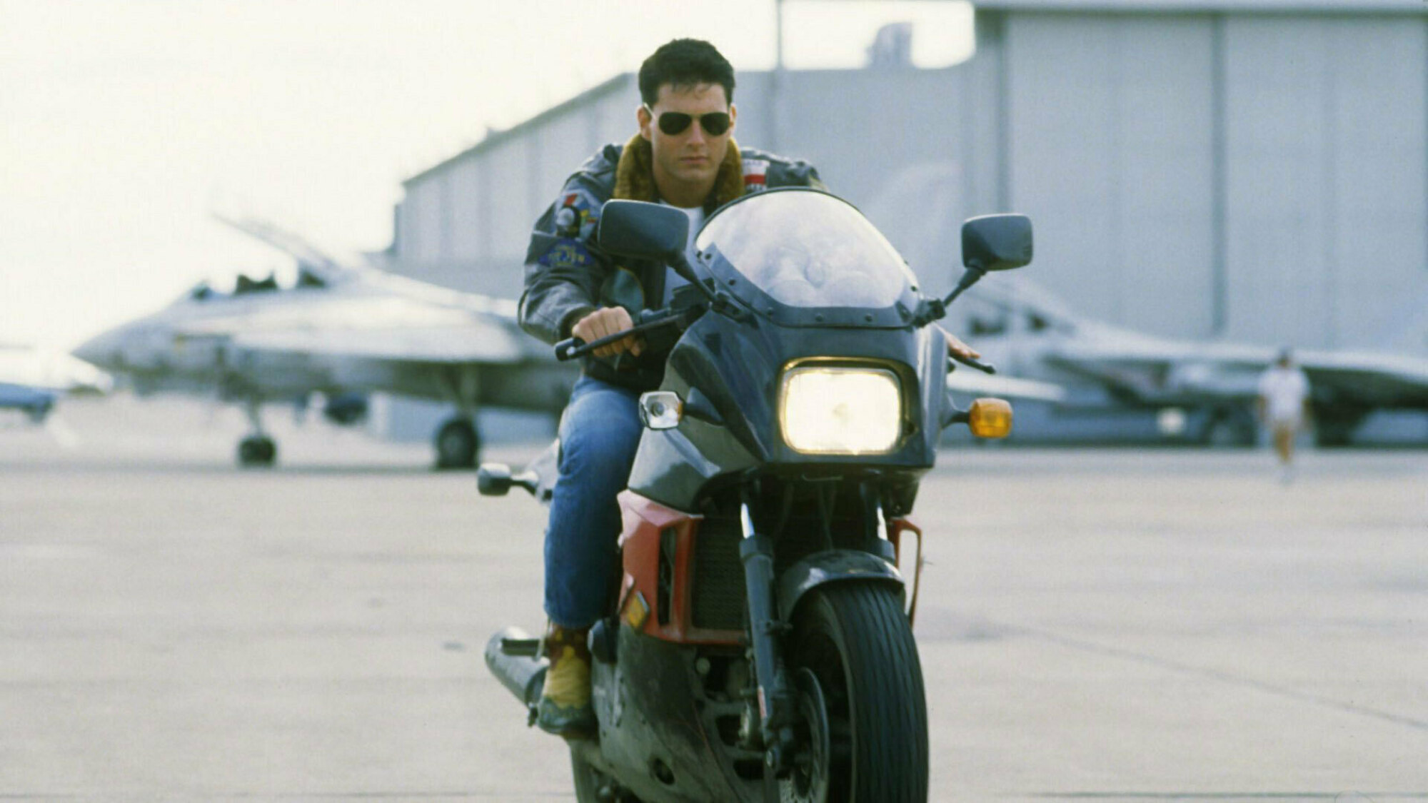 Un homme sur une moto, portant une veste en cuir et des lunettes de soleil aviateur. 