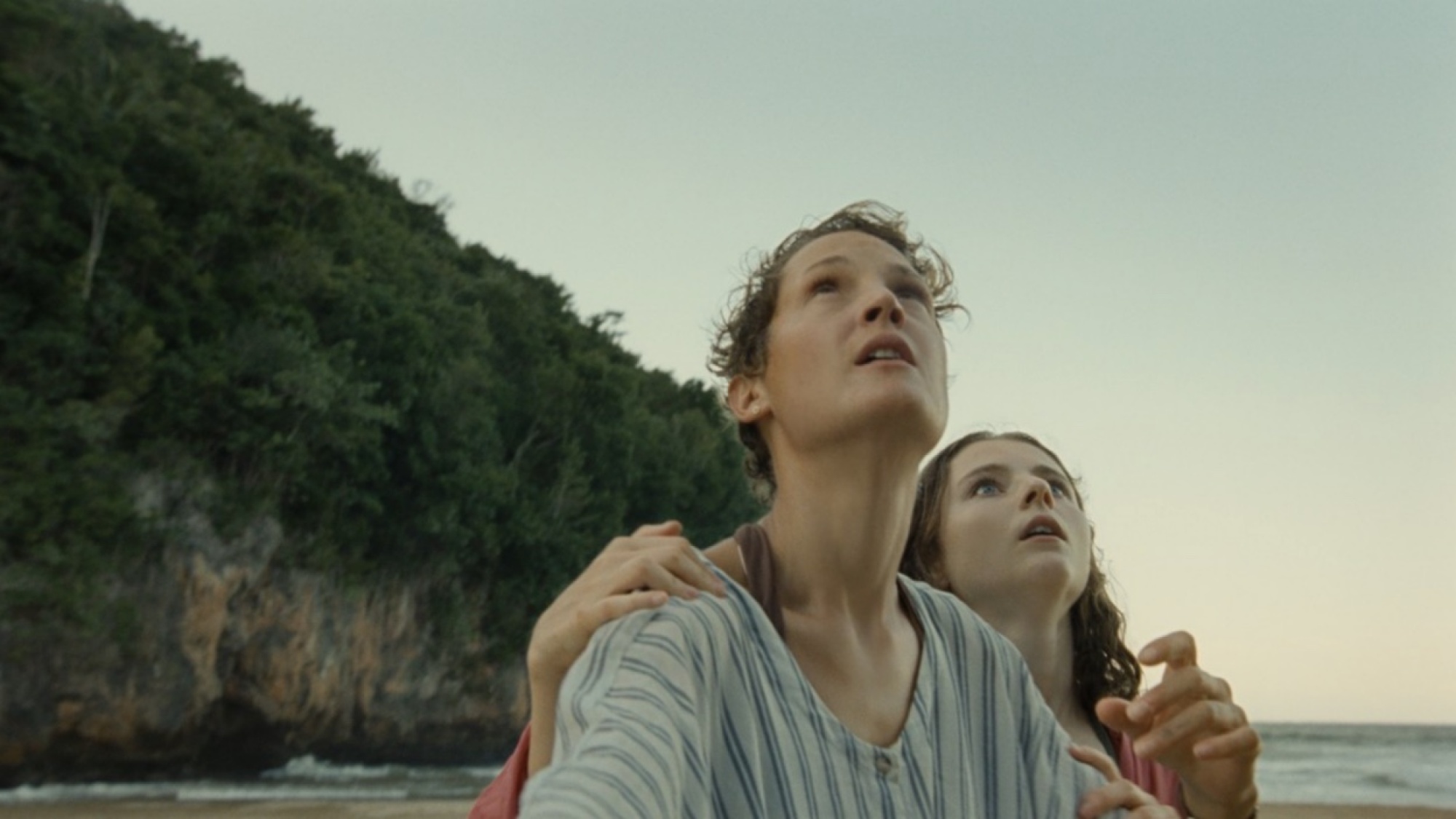 Une femme aux cheveux courts serrant une jeune fille sur une plage. 