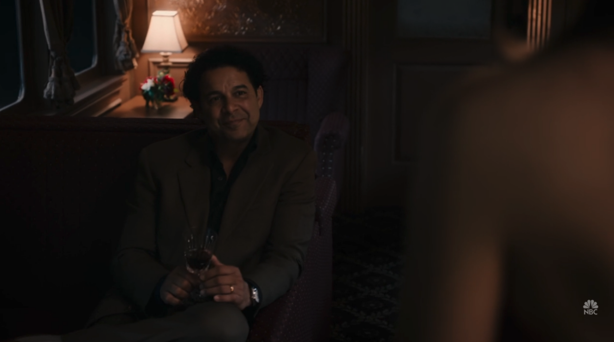 Un homme (Jon Huertas) assis dans un train tenant un verre de vin.