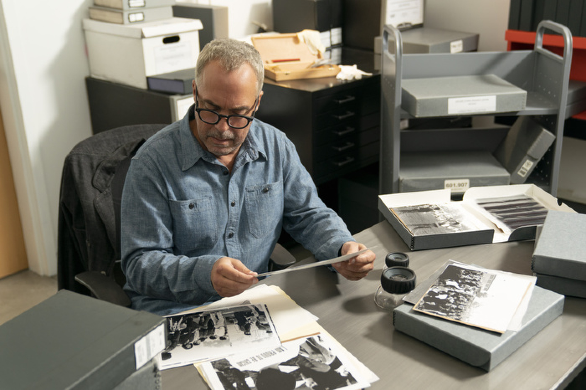 Un homme est assis à un bureau et regarde des photographies en noir et blanc.