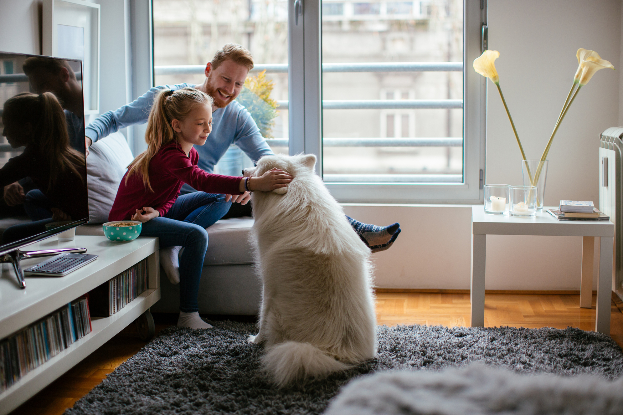 Image d'un père et d'une jeune fille assis sur une chaise en train de caresser un chien qui est assis et leur fait face.