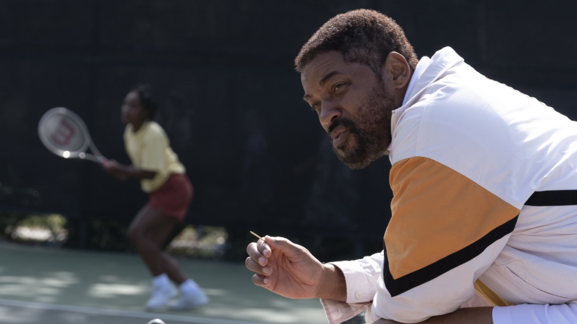 Un homme est assis en marge d'un court de tennis tandis qu'une jeune fille s'entraîne à l'arrière.