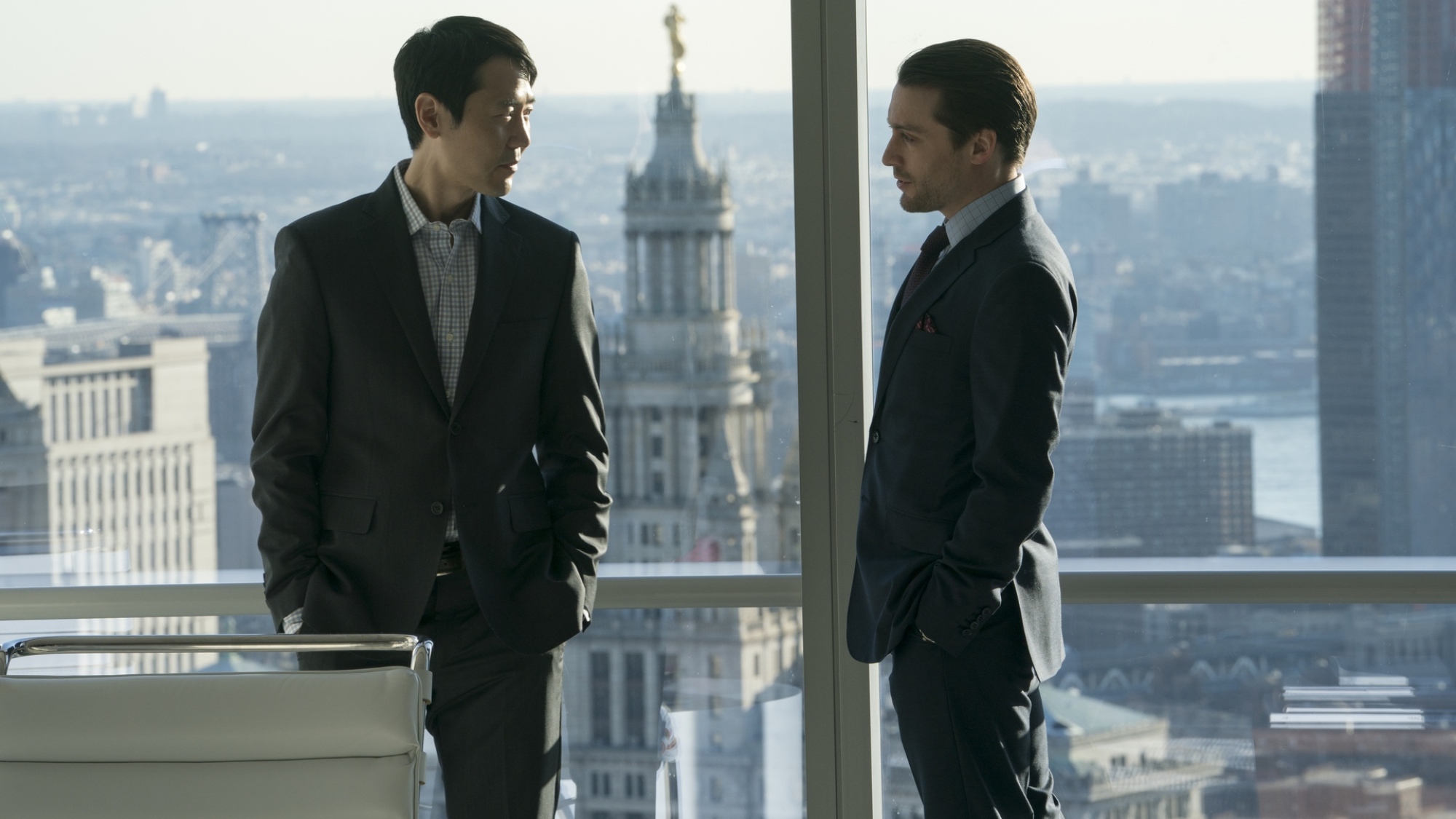 Deux hommes en costume se tiennent dans un bureau, devant une grande baie vitrée, et se regardent.