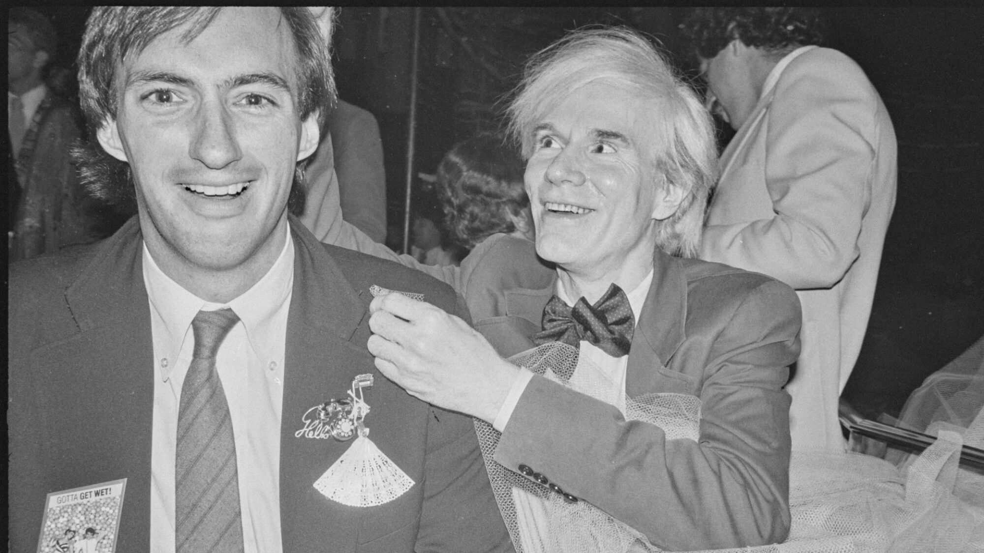Une photo en noir et blanc de deux hommes en costume ;  l'un est Andy Warhol. 