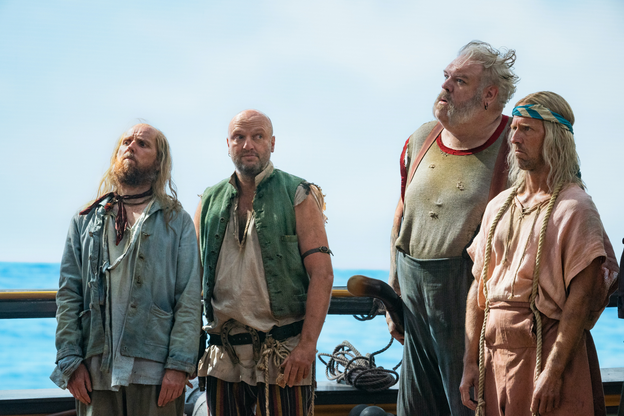 Quatre pirates en haillons se tiennent sur le pont de leur navire avec l'océan derrière eux.
