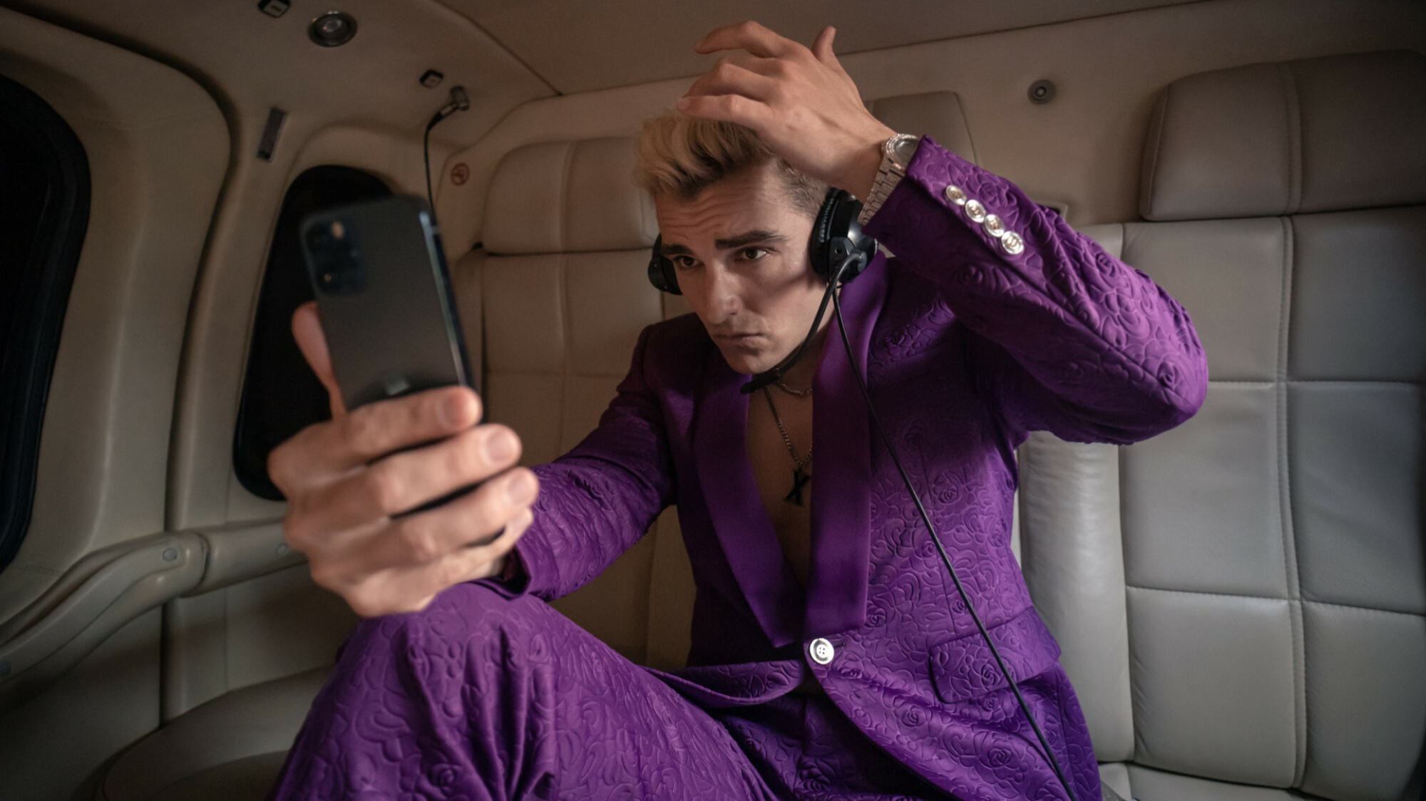 Un homme en costume violet avec des cheveux blonds décolorés se vérifie dans son téléphone;  un alambic de 
