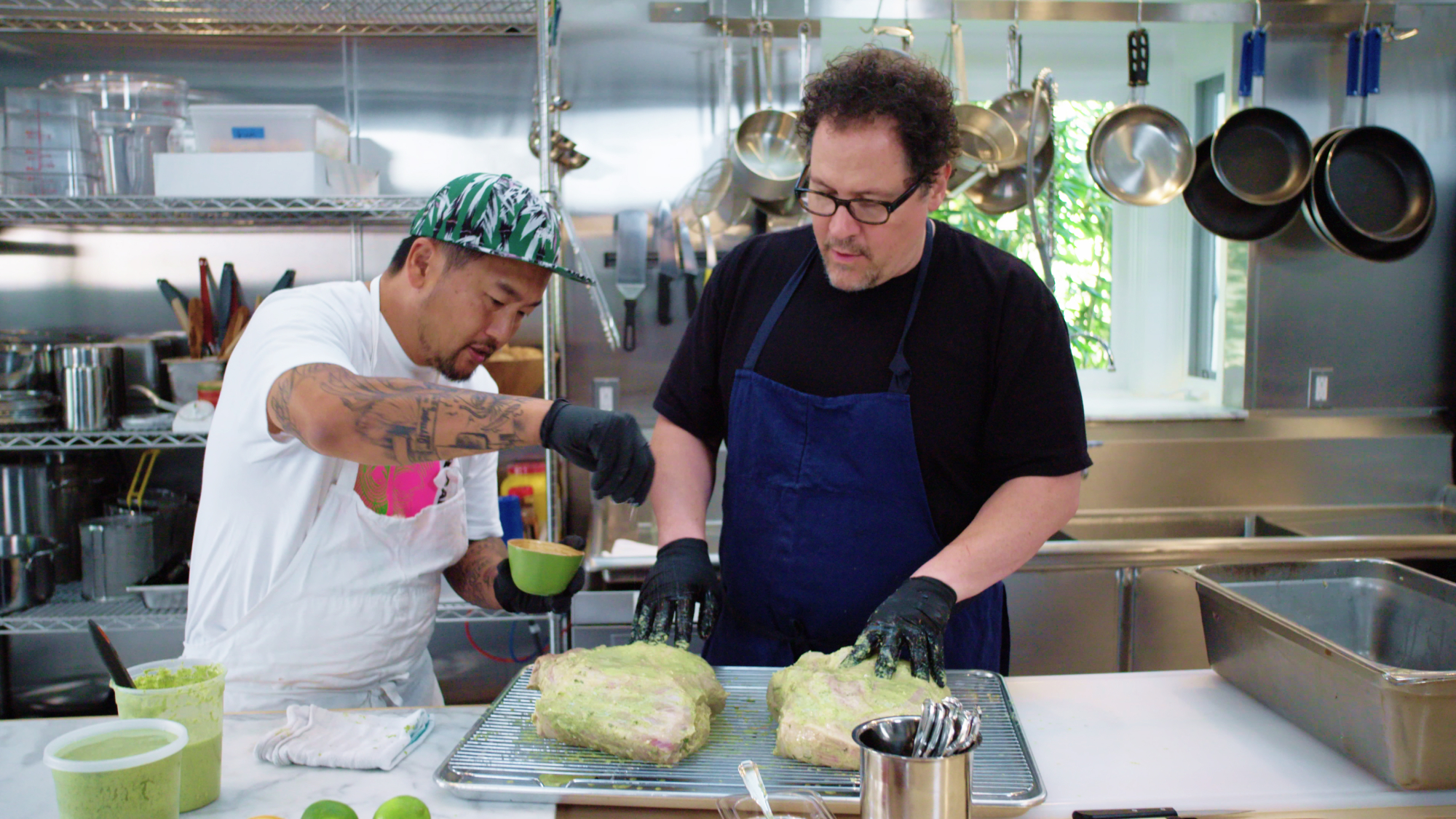 Deux hommes, les hôtes Roy Choi et Jon Favreau, se tiennent côte à côte derrière un comptoir de cuisine.  Ils assaisonnent deux gros morceaux de viande.