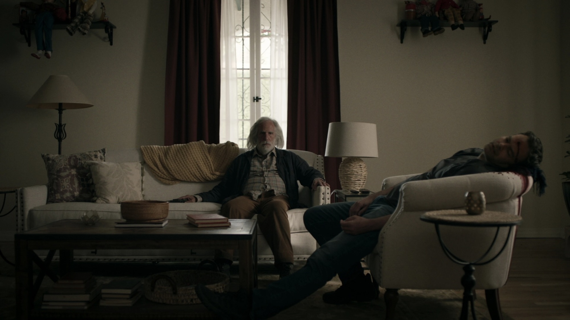 Gene Cousineau est assis sur un canapé, tandis que le cadavre de Barry Berkman avec une balle dans la tête repose sur une chaise à proximité.