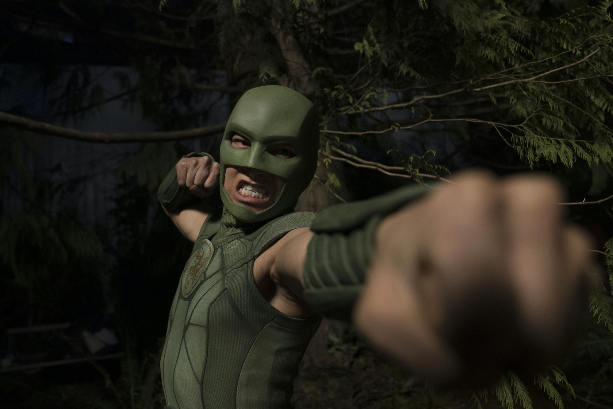 Une personne portant un masque et un body verts, prête au combat ;  un alambic de 