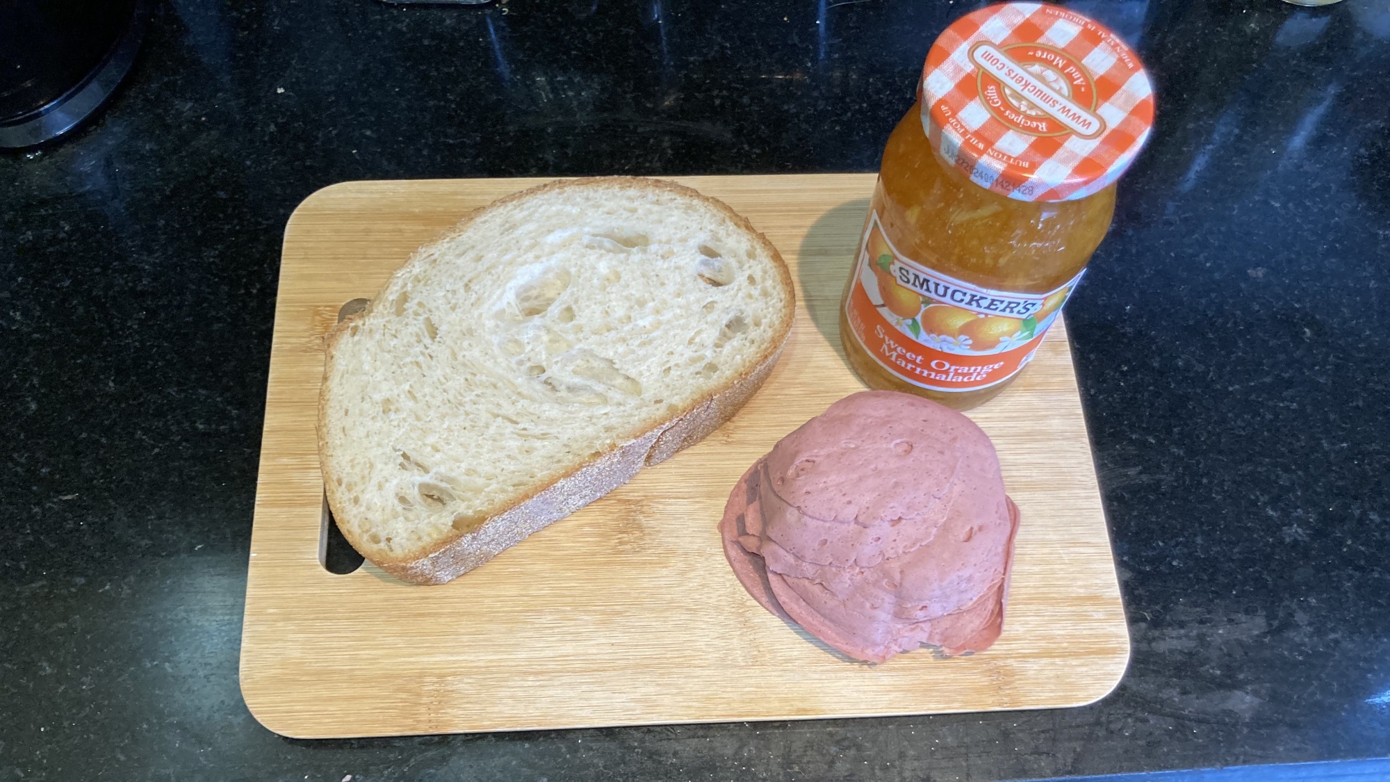 Deux tranches de pain, un pot de marmelade et un tas de saucisses de foie en tranches sont assis sur une planche à découper en bois sur un comptoir noir.