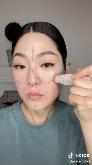 Une femme asiatique aux cheveux noirs regarde la caméra et applique le contour à la joue avec un filtre superposé.