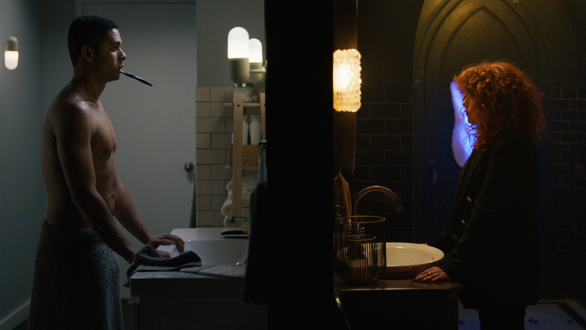 Images côte à côte d'un homme et d'une femme face aux miroirs de deux salles de bains respectives ;  son nu, gris et brillamment éclairé tandis que le sien est éclectique et sombre avec une lumière bleue étrange;  encore de 