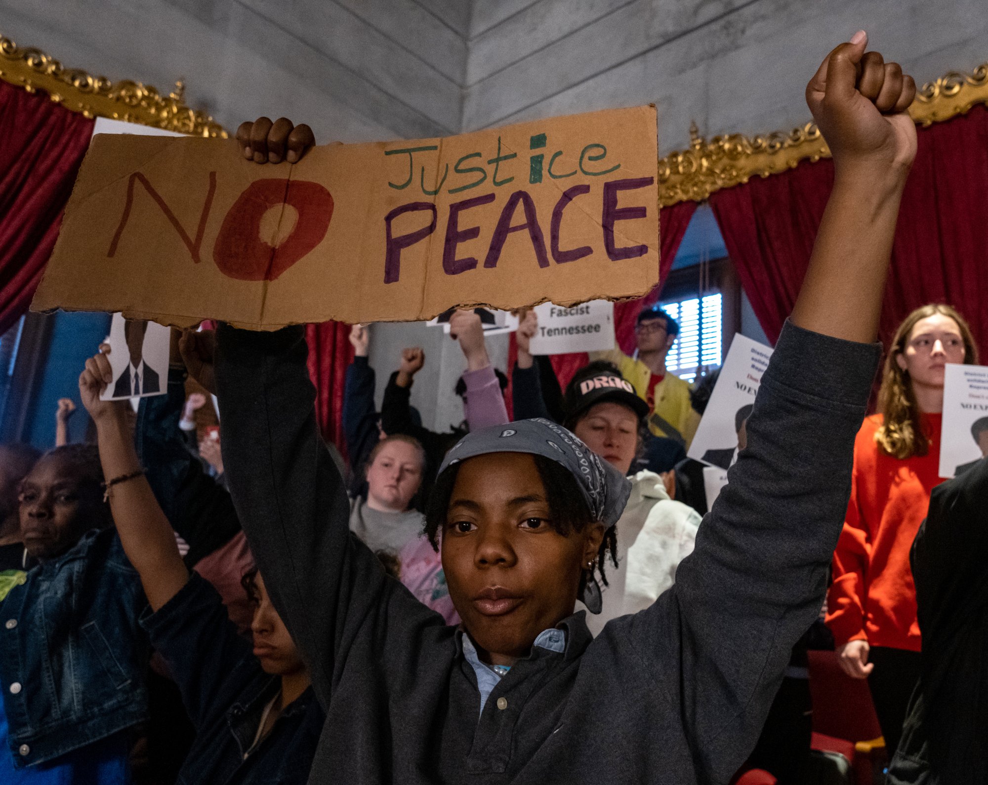 Un jeune se tient debout, le poing gauche en l'air et la main droite tenant une pancarte indiquant 