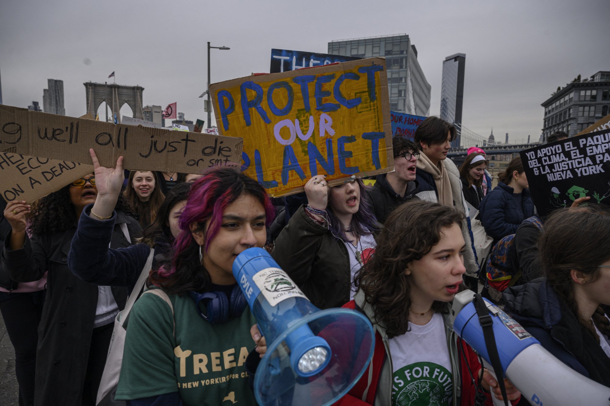 Une grande foule de manifestants traverse le pont de Brooklyn brandissant des pancartes et des mégaphones.  Un signe lit, 