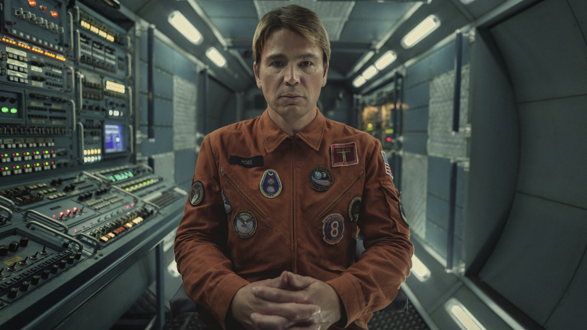 Un homme vêtu d'une combinaison d'astronaute orange regarde fixement la caméra depuis l'intérieur d'une salle de contrôle.