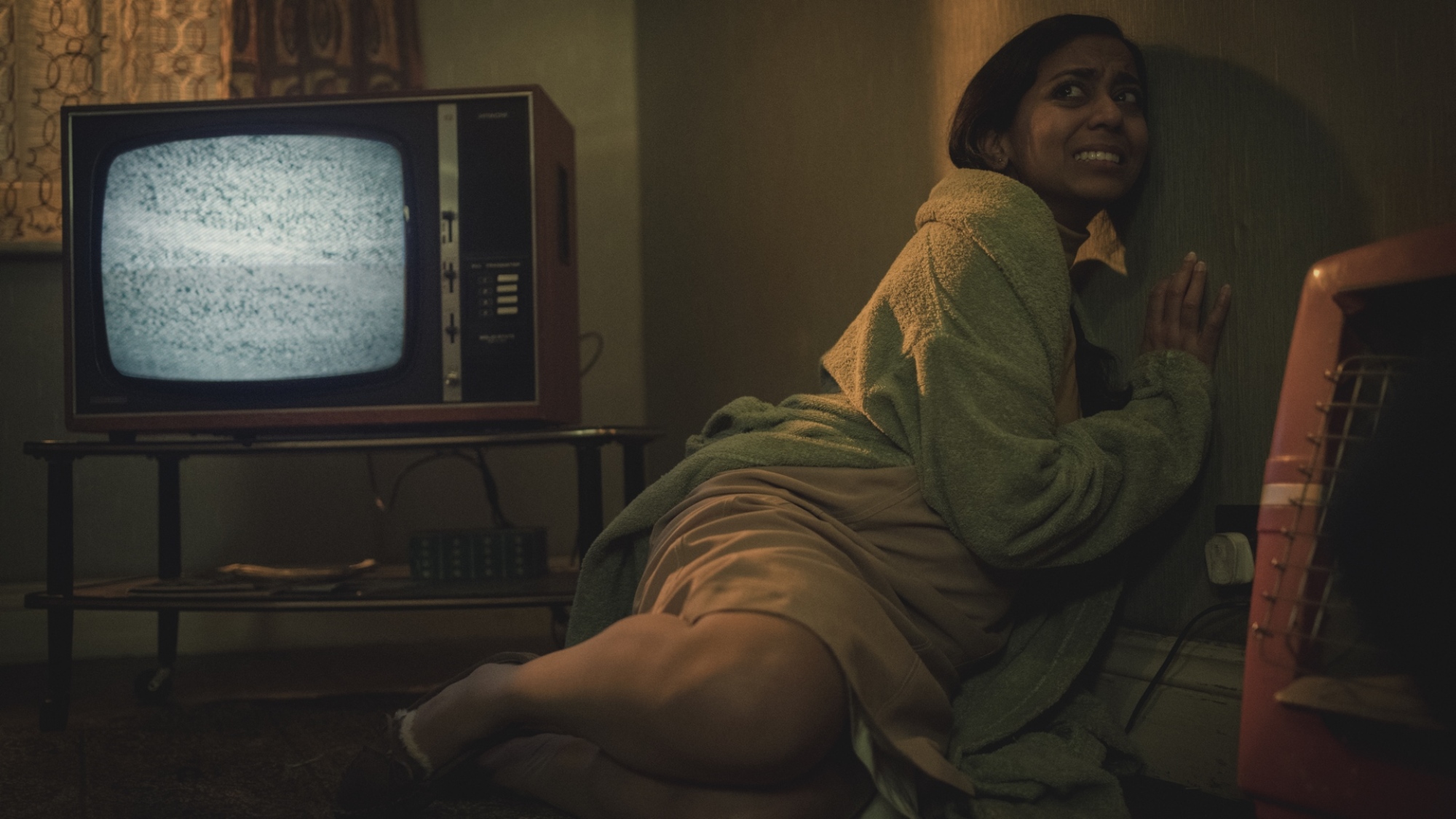 Une femme en tenue des années 70 se recroqueville contre le mur tandis qu'un écran de télévision à côté d'elle affiche des parasites.