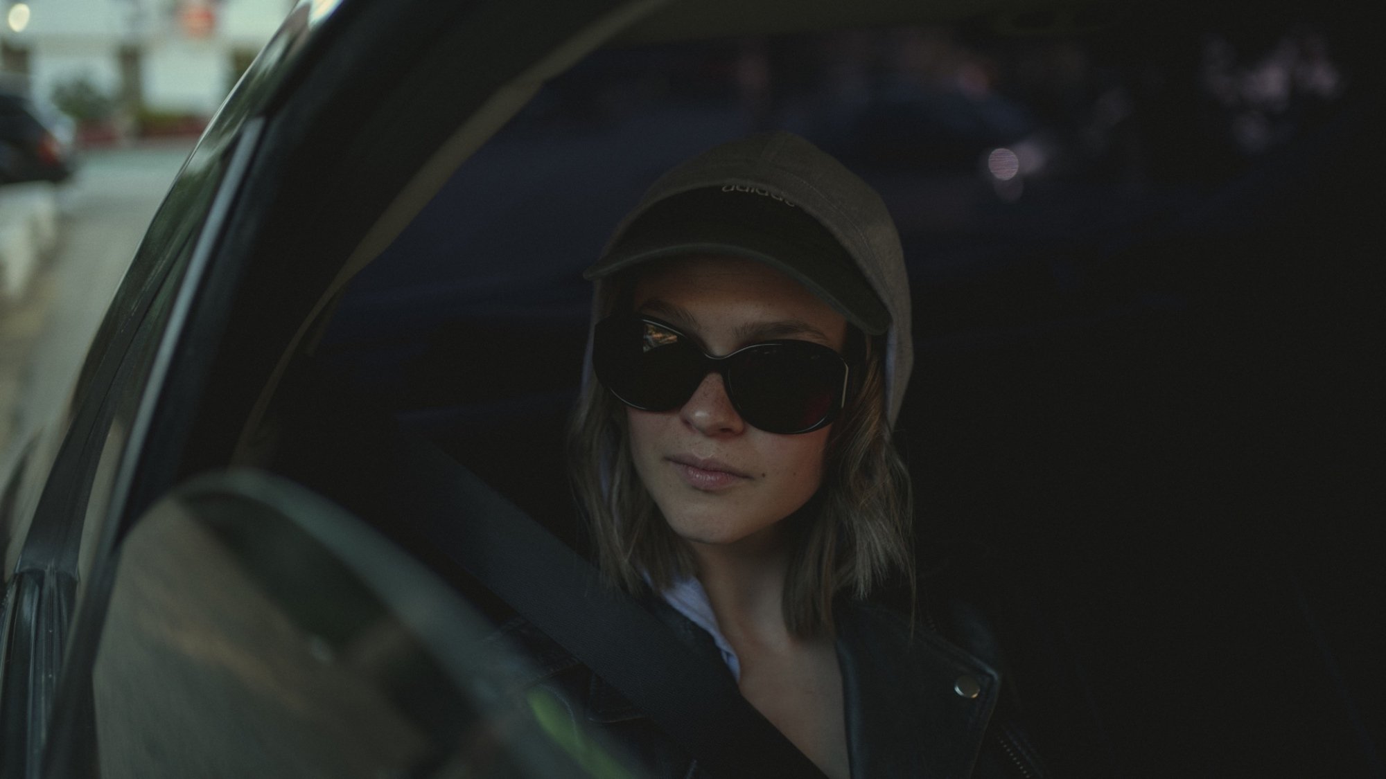 Une femme portant des lunettes de soleil et un sweat à capuche est assise sur le siège arrière d'une voiture avec la fenêtre baissée.