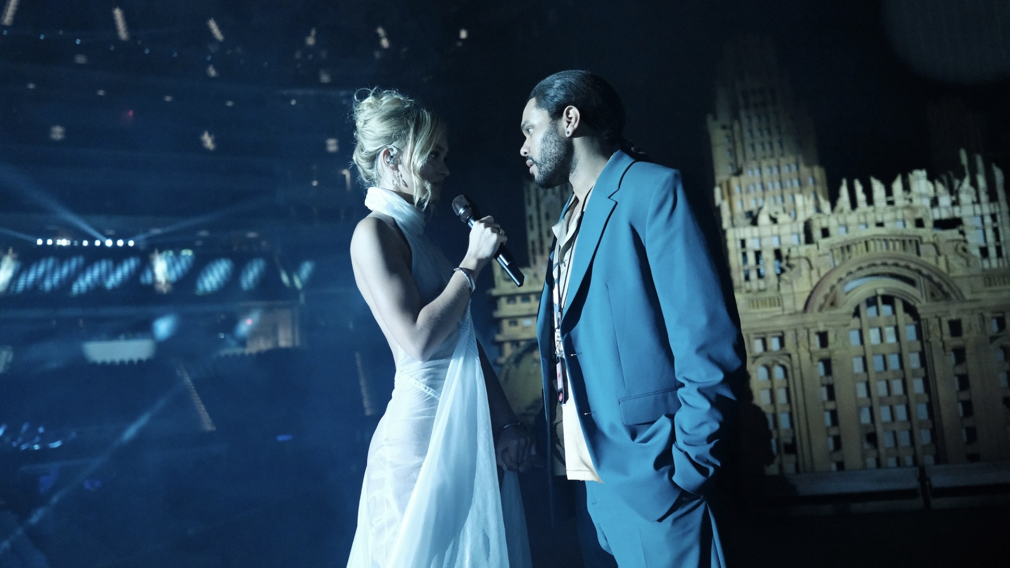 Une femme en robe blanche tenant un microphone sur scène dans un stade parle à un homme en costume bleu.