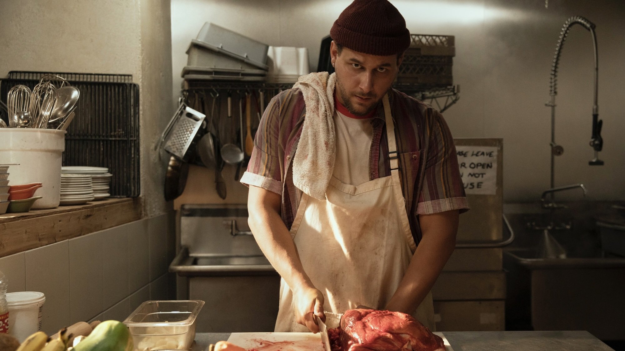 Un homme portant un tablier et un bonnet marron coupe de la viande avec un couperet dans la cuisine d'un restaurant.