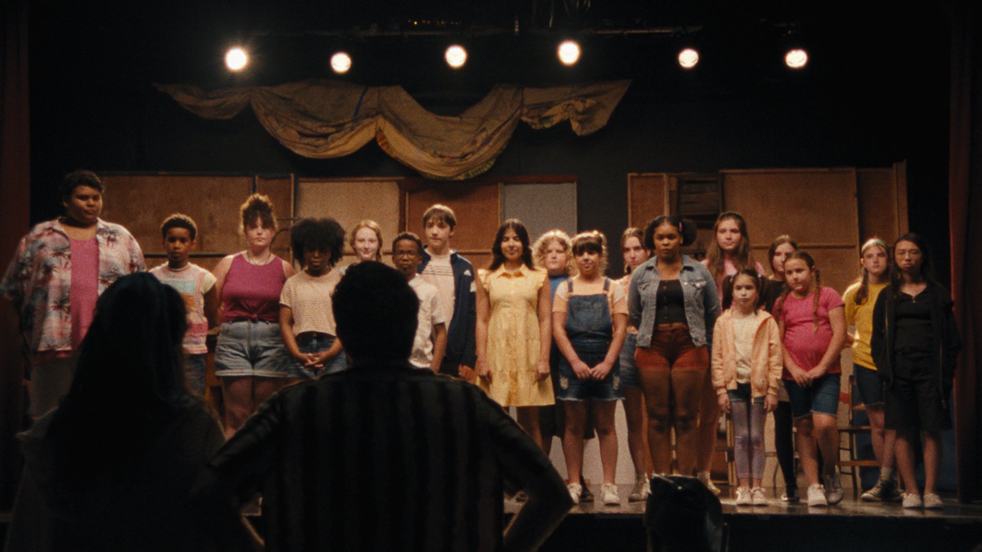 Un groupe d'enfants se tient sur scène tandis qu'un homme et une femme silhouettes regardent.