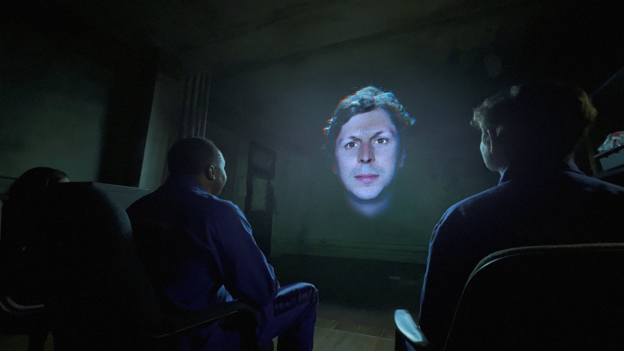 Une image du visage de Michael Cera rayonnant à travers un écran vers trois personnes assises dans une pièce sombre.