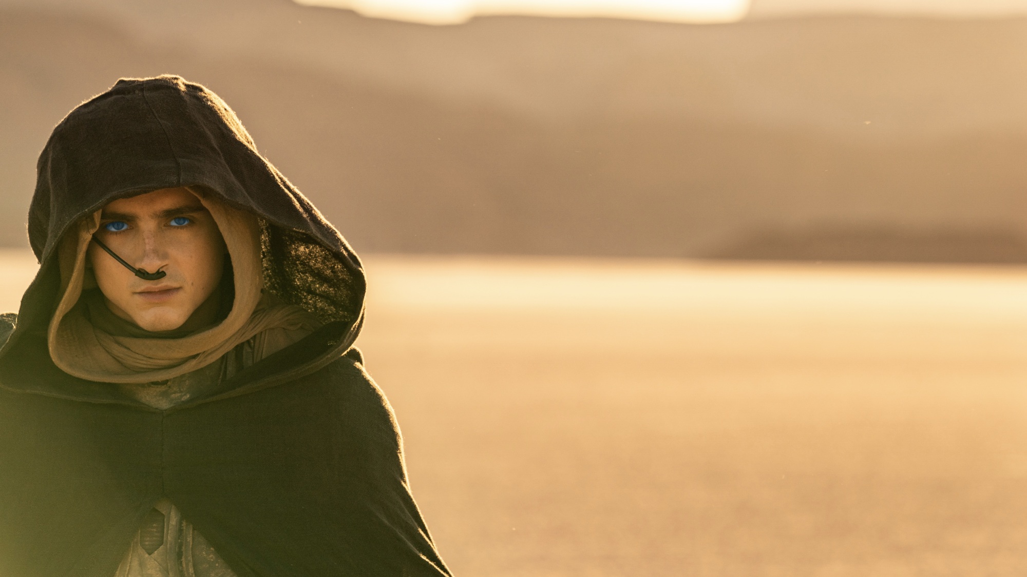 Un jeune homme aux yeux bleu vif marche dans le désert vêtu d'une cape sombre à capuche.