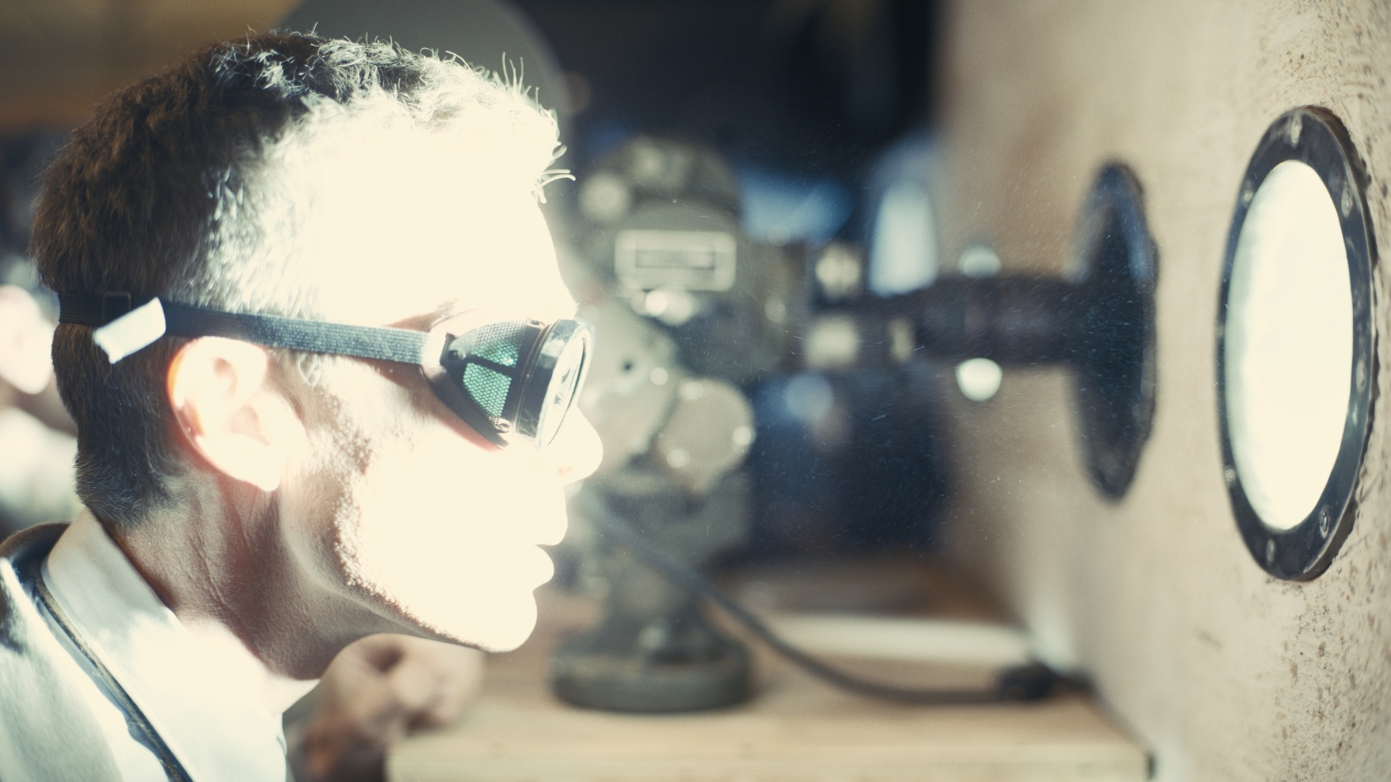 Un homme portant des lunettes regarde par une fenêtre circulaire, baignée de la lumière blanche brillante d'une explosion.