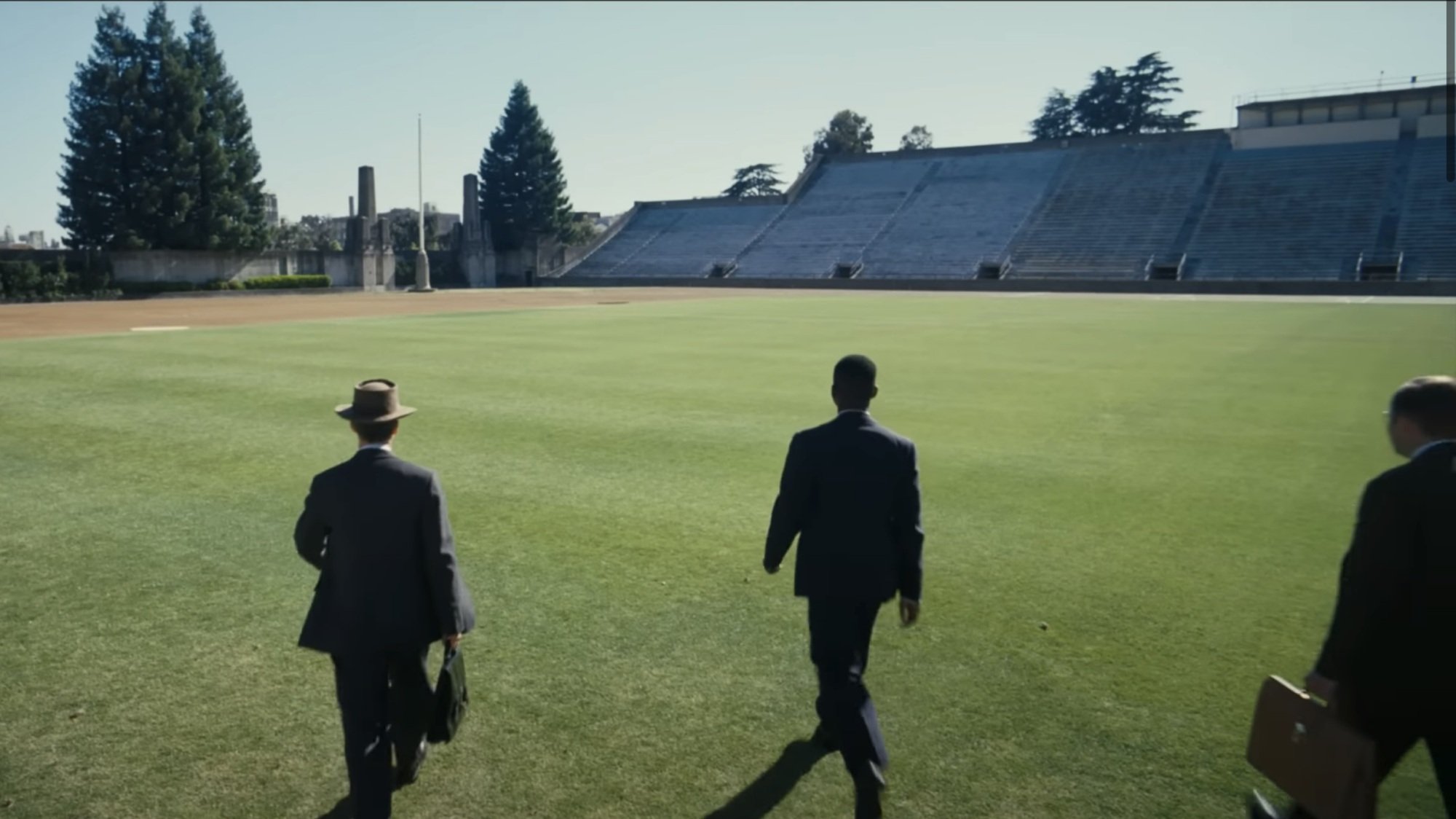 Trois hommes en costume noir traversent un terrain de football universitaire.