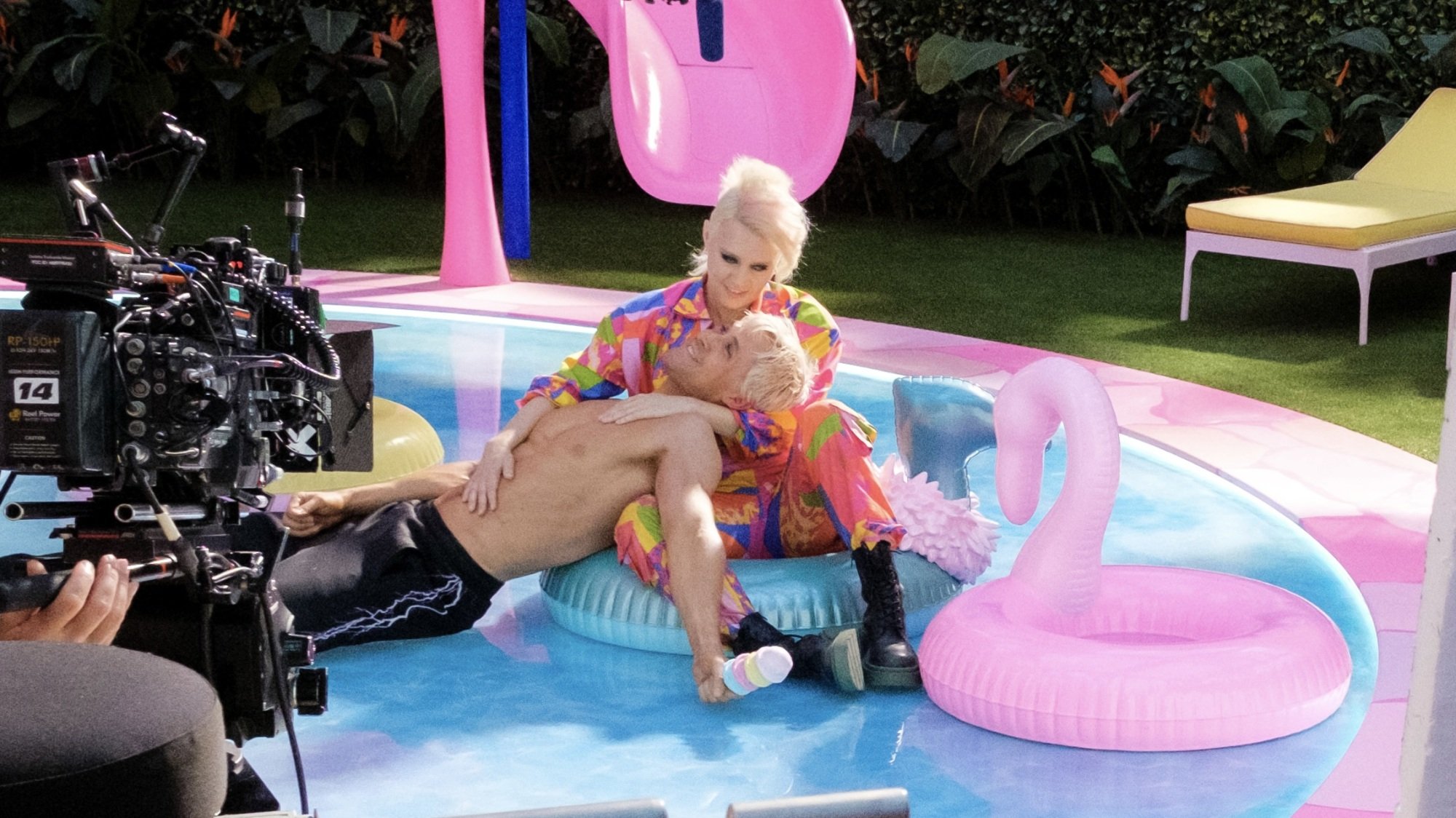 Ryan Gosling dans le rôle de Ken est allongé torse nu dans Kate McKinnon sur les genoux de Barbie.  Les deux flottent sur une fausse piscine bleue.