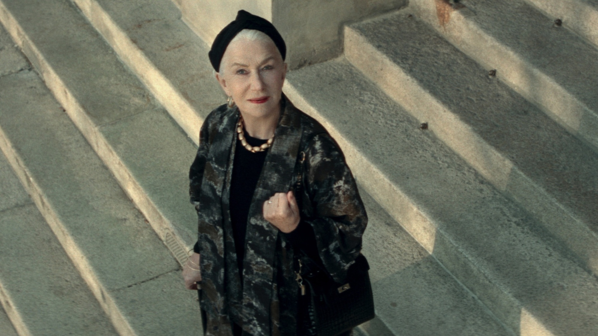 Une femme vêtue d'un long manteau noir se tient dehors sur des marches de pierre, souriant au ciel.
