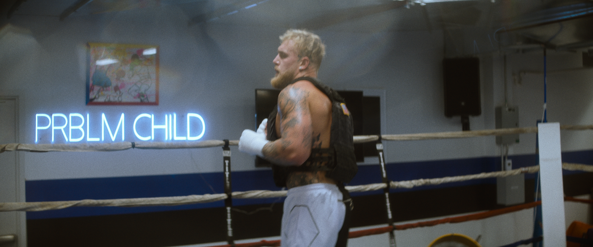 Jake Paul sur le ring de boxe au gymnase.  Derrière lui, une enseigne au néon indique 