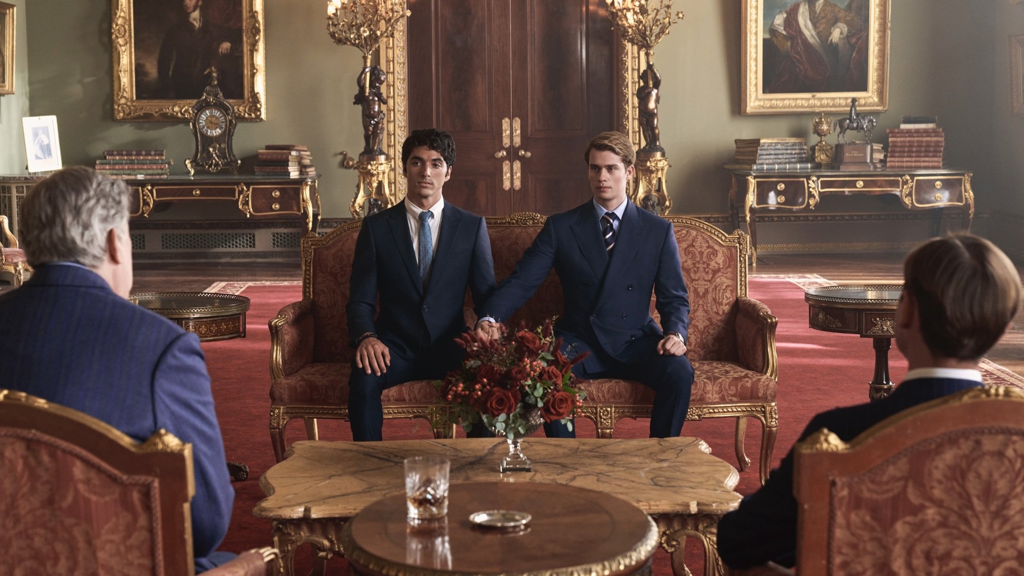 Deux hommes en costume se tiennent la main assis sur un canapé rouge à Buckingham Palace.