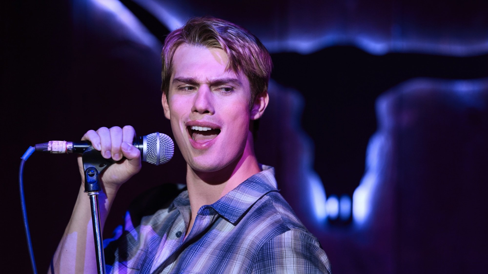 Un homme blond vêtu d'une chemise à carreaux bleus chante un karaoké passionné dans un bar éclairé en violet.