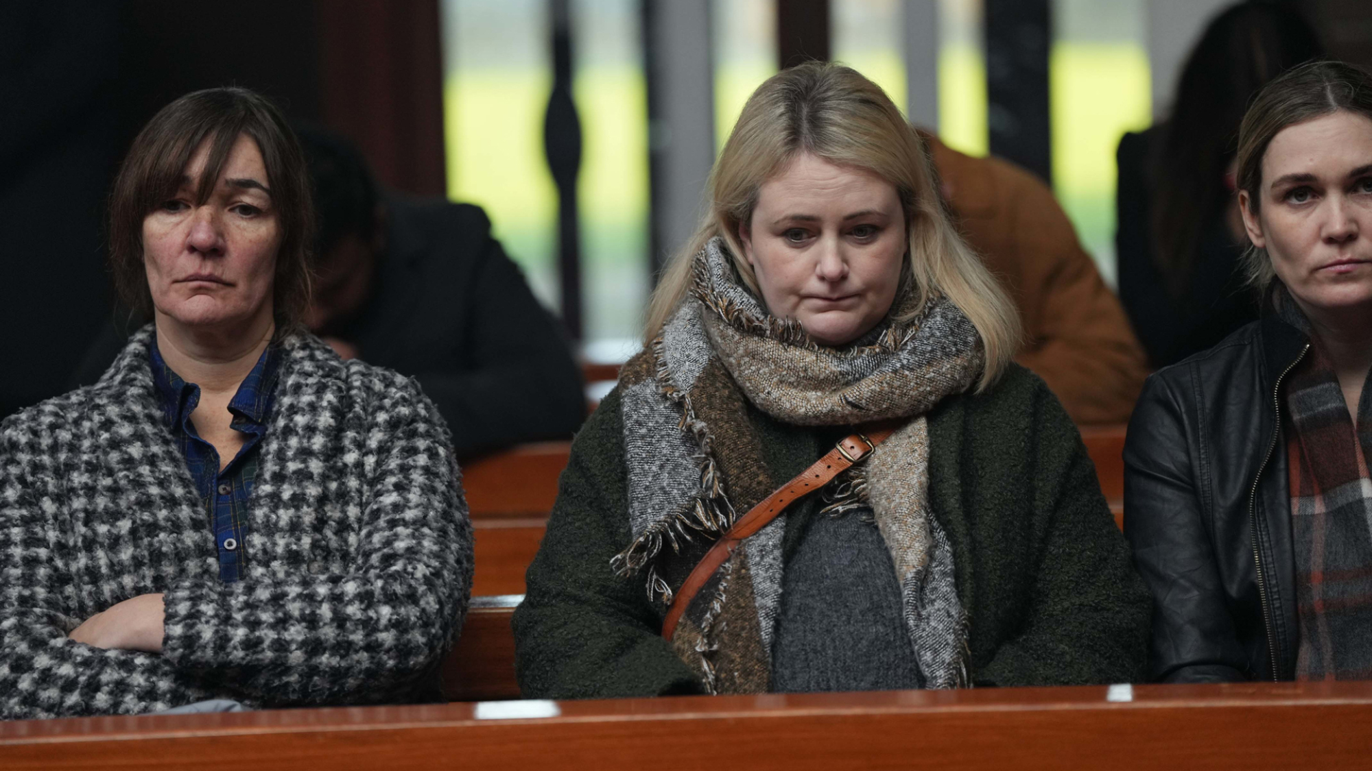 Trois femmes à l’air maussade sur un banc d’église, portant des vêtements chauds de couleur terne.