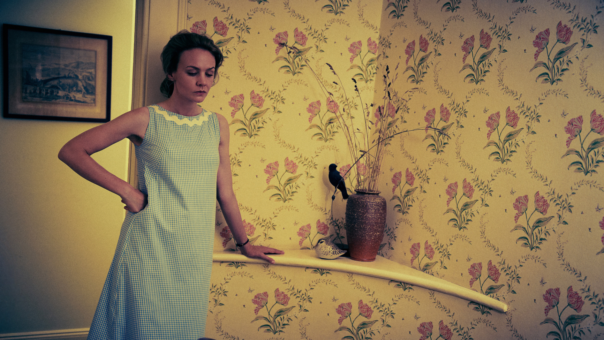 Carey Mulligan dans le rôle de Felicia Montealegre dans le film "Maestro", debout, sombre dans un décor domestique avec du papier peint jaune.