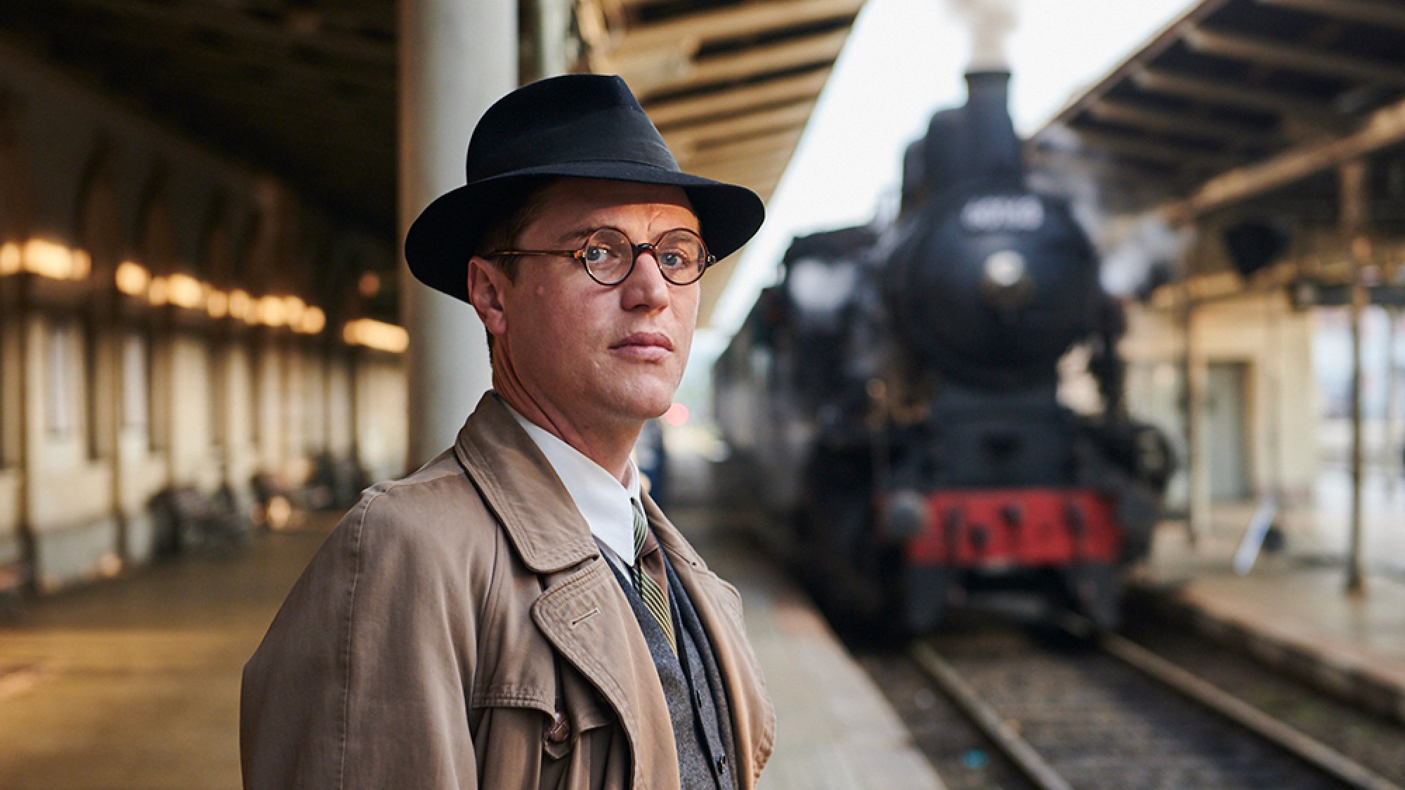 Un jeune homme portant des lunettes se tient sur un quai de train avec un train à vapeur en arrière-plan.